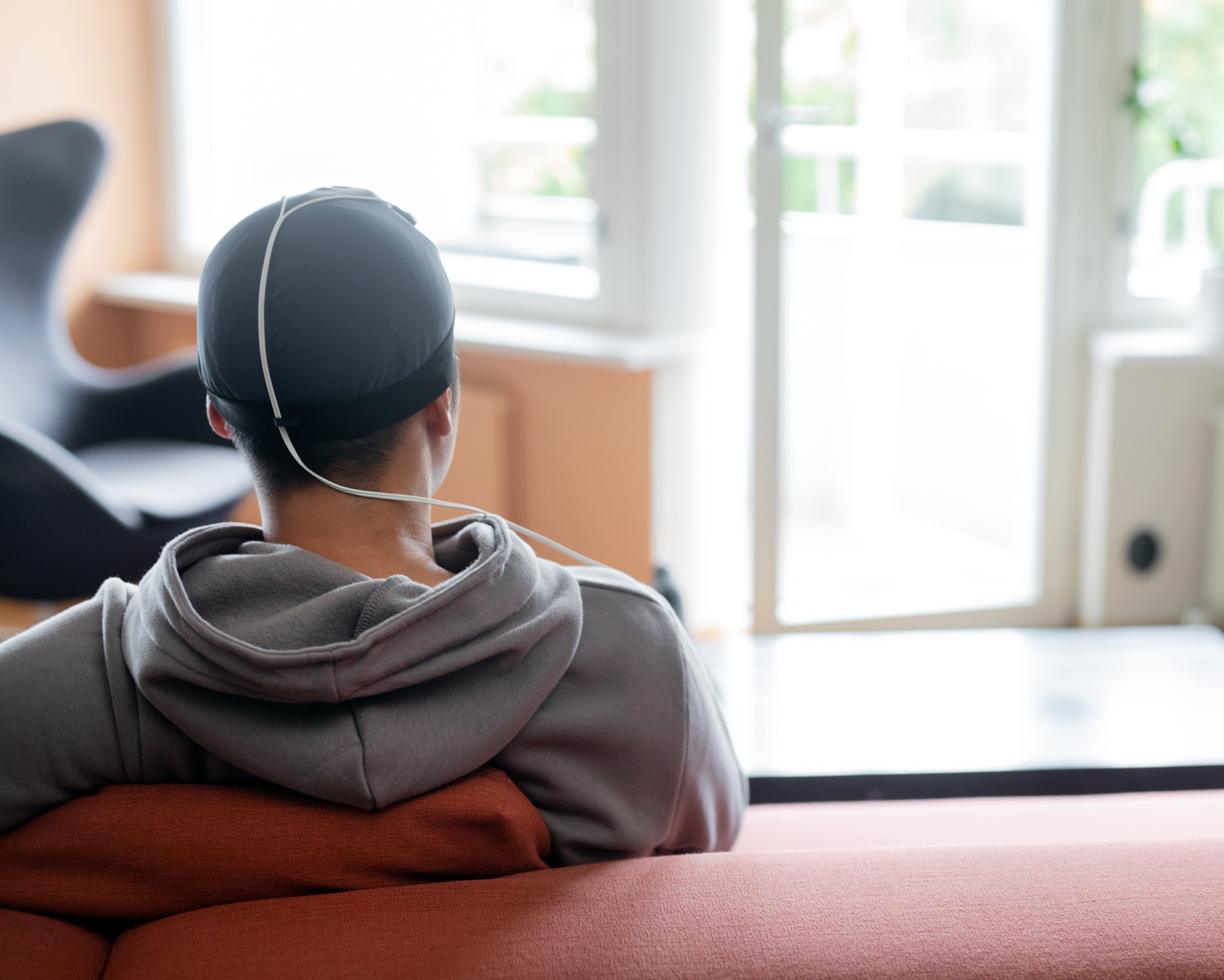 A person seen from behind sitting on a terracotta-colored couch in a bright room, wearing a Sooma ComfoCap with a white cord attached.